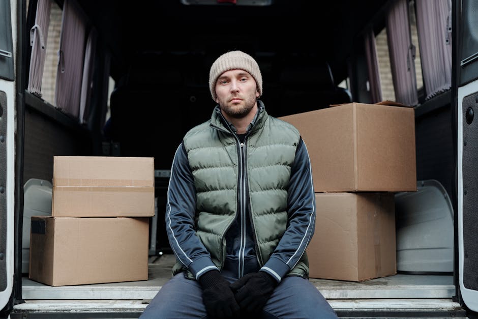 A young man with a serious expression sitting inside the open rear of a van used for house removals, with several cardboard boxes of various sizes stacked around him. He is wearing a beige knit beanie, a green puffer vest over a dark long-sleeved sports jacket, and black gloves. The van's interior is visible behind him, showing more packed boxes and moving equipment. Outside the van, a paved area and a building exterior can be seen, indicating a loading or unloading environment for home relocation services. The image captures the process of furniture transport and packing in a professional moving context, with the man representing the logistical side of house removals by [COMPANY_NAME], highlighting the importance of careful loading and transport during a move.