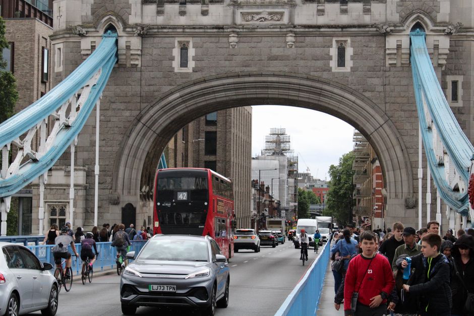A busy urban street scene during daytime shows the Tower Bridge in London with its iconic blue suspension cables and stone archway. In the foreground, pedestrians, including children and adults, are walking along the sidewalk and crossing the road, some carrying bags or engaged in conversation. Several bicycles and cyclists are visible among the pedestrians. Vehicles, including a red double-decker bus, a silver car, and other cars, are moving through the intersection underneath the bridge. The street is lined with shop fronts and buildings, with scaffolding and construction work visible on some structures in the distance. The scene features overcast lighting, typical of London's weather, and captures a moment of active home relocation or moving process, possibly involving the transportation of furniture and boxes, as part of a professional removals service provided by Man and Van London.