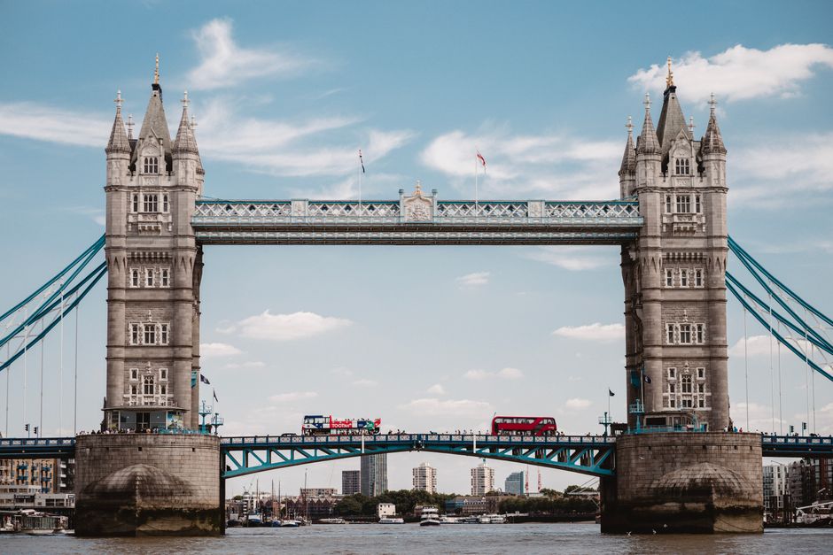 A view of Tower Bridge in London during daylight, with its Gothic-style towers and suspension cables visible. In the foreground, a man and van company, Man and Van London, is engaged in a home relocation process, with a van parked nearby. Inside the property, several cardboard boxes, wrapped furniture, and household appliances are being prepared for transit. Moving blankets and plastic wrap are used to secure and protect the items, while a trolley and straps are visible for lifting and stability. Two people are seen carrying a large cardboard box towards the vehicle, with other boxes and packing materials arranged close to the entrance, indicating an active loading process. The scene captures the logistical aspect of furniture transport and packing and moving services relevant to house removals near Tower Bridge, with an emphasis on careful handling and organized loading activities on a clear, bright day.