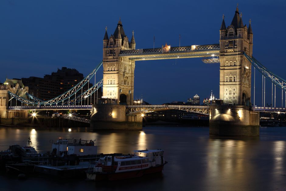 A busy urban street scene during daytime shows the Tower Bridge in London with its iconic blue suspension cables and stone archway. In the foreground, pedestrians, including children and adults, are walking along the sidewalk and crossing the road, some carrying bags or engaged in conversation. Several bicycles and cyclists are visible among the pedestrians. Vehicles, including a red double-decker bus, a silver car, and other cars, are moving through the intersection underneath the bridge. The street is lined with shop fronts and buildings, with scaffolding and construction work visible on some structures in the distance. The scene features overcast lighting, typical of London's weather, and captures a moment of active home relocation or moving process, possibly involving the transportation of furniture and boxes, as part of a professional removals service provided by Man and Van London.